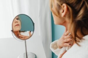 Woman in a bathrobe applying skincare while looking in the mirror.